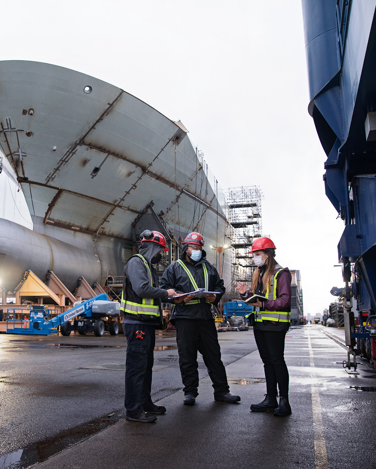 Three Seaspan employees wearing red hard hats and safety vests talking in front of a cargo ship.